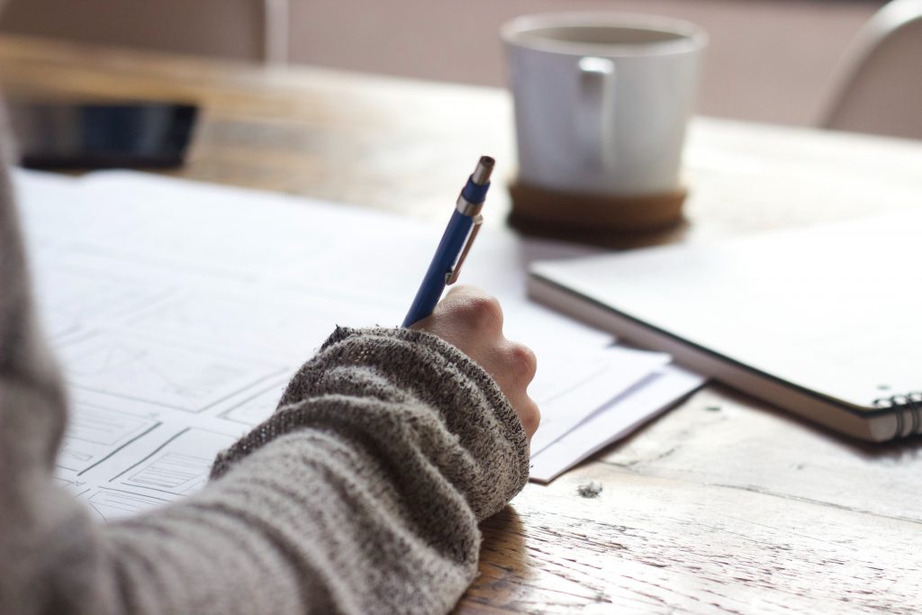 A close up of a desk with a persons hand holding a pen writing on a piece of paper. There is a mug in the background on the desk