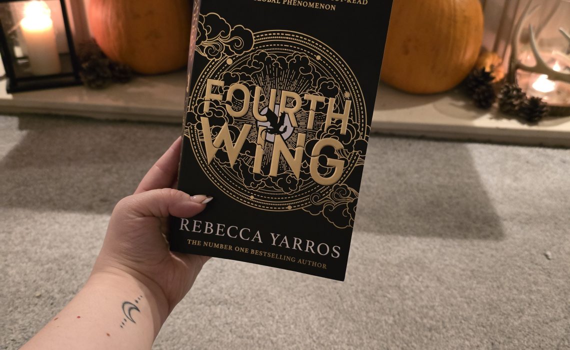 Fourth Wing by Rebecca Yarros book being held up in front of a cosy fireplace with pumpkins